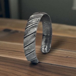 May include: A Damascus steel cuff bracelet with a unique pattern of swirling lines and dark accents. The open-ended design features a polished finish, showcasing the intricate metalwork. This handcrafted piece is a statement accessory.