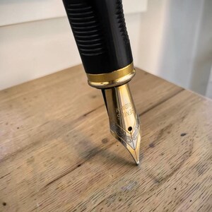Op de afbeelding: Een vulpen met een zwart geribbeld gripgedeelte en een goudkleurige metalen ring. De penpunt is goudkleurig met ingewikkelde zilverkleurige gravures en de tekst GENIUS IRIDIUM is duidelijk zichtbaar op het oppervlak van de metalen penpunt.