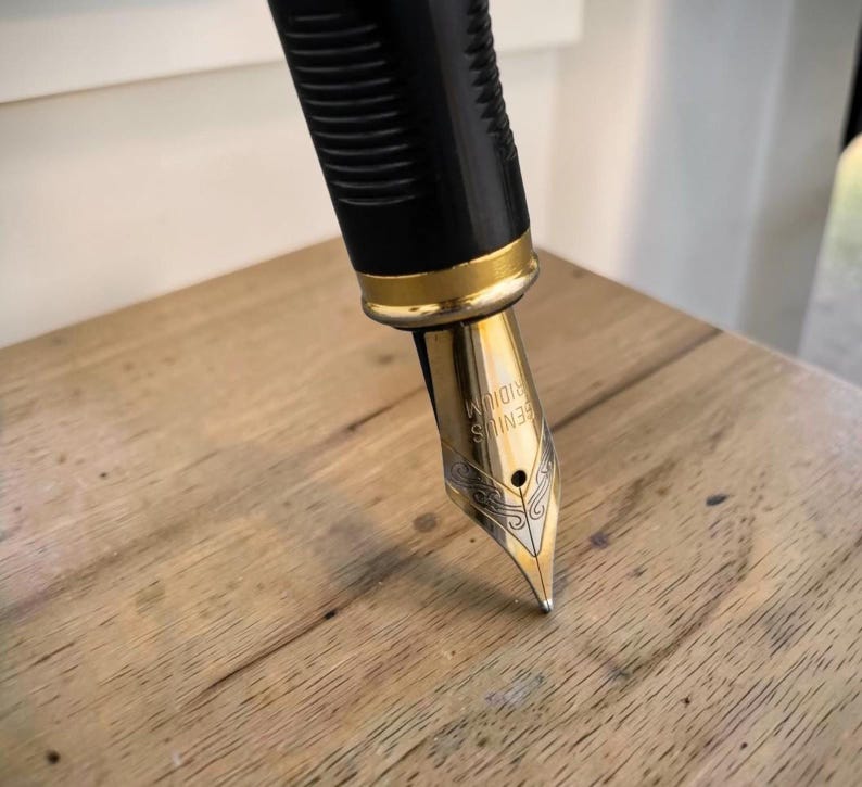 Op de afbeelding: Een vulpen met een zwart geribbeld gripgedeelte en een goudkleurige metalen ring. De penpunt is goudkleurig met ingewikkelde zilverkleurige gravures en de tekst GENIUS IRIDIUM is duidelijk zichtbaar op het oppervlak van de metalen penpunt.