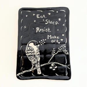 May include: A rectangular black ceramic tray with a glossy finish featuring a white hand-painted illustration of a bird perched on a branch. The text "Eat. Sleep. Resist. Make art." is written in white, surrounded by small stars and hearts.