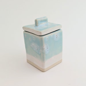 May include: A square ceramic canister with a matching lid, featuring a light blue glaze with white crystalline patterns on the upper portion and a solid white base. The container has a rectangular handle on top of the lid and a smooth finish.