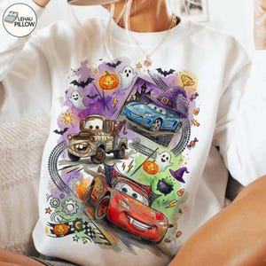 May include: A white long-sleeve sweatshirt featuring a colorful Halloween-themed graphic with cartoon cars, including a red race car, a brown tow truck, and a blue car wearing a witch hat, surrounded by ghosts, pumpkins, bats, and candy.