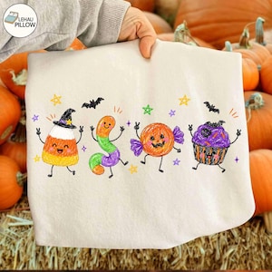 May include: A white sweatshirt featuring a colourful crayon-style illustration of four Halloween characters: a candy corn wearing a witch hat, a gummy worm, a jack-o'-lantern candy, and a cupcake with purple frosting, all surrounded by small stars.