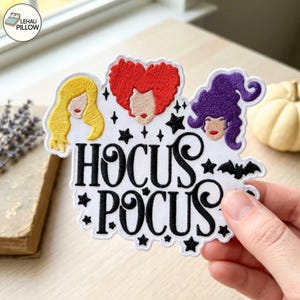 May include: A white embroidered patch featuring the text HOCUS POCUS in black script. Above the text are three stylised hair silhouettes in yellow, red, and purple, accented by small black stars and a bat silhouette on a white fabric background.