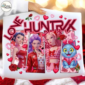Könnte beinhalten: Weißes Kissen mit dem pinkfarbenen Text "LOVE HUNTRX". Das Design zeigt Cartoon-Illustrationen von drei Frauen und einer blauen Katze, umgeben von Herzen und Rosen. Das Kissen wird von einer Person gehalten.