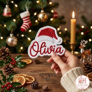 May include: A white embroidered patch featuring the name Olivia in red script lettering, topped with a red and white Santa hat design. The patch has a white border and is held up against a festive holiday background with lights.