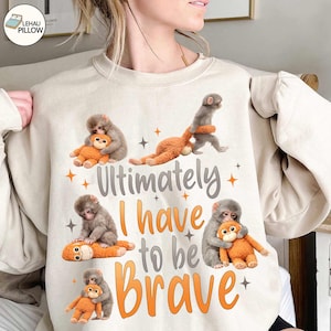 May include: Beige sweatshirt featuring a graphic of several gray monkeys hugging orange stuffed animals. The text "Ultimately I have to be Brave" is printed in gray and orange. The design includes orange star accents.