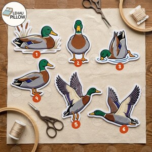 May include: Six embroidered mallard duck patches in various poses, numbered one through six. The designs feature green heads, brown chests, and grey wings with blue accents. The patches are arranged on a light fabric surface with scissors and thread.