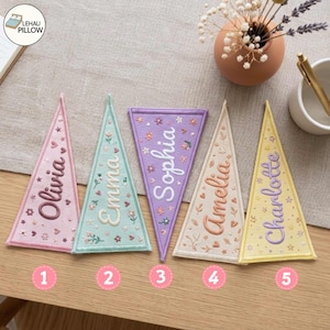 Könnte beinhalten: Fünf dreieckige Filzwimpel in Pastellrosa, Mintgrün, Lavendel, Creme und Hellgelb. Jeder Wimpel ist mit gestickten Blumenmotiven, Herzen und einem Namen versehen: Olivia, Emma, Sophia, Amelia oder Charlotte, dazu nummerierte rosa Kreise.