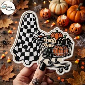 May include: An embroidered patch featuring a ghost with a black and white chequered pattern pushing a shopping cart filled with orange, white, and black pumpkins. The patch has a white border and is shaped to follow the outline of the ghost and cart.