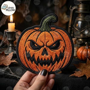 May include: A round embroidered patch featuring a bright orange pumpkin with a dark green stem. The pumpkin has a menacing jack-o'-lantern face with black stitched eyes, a triangular nose, and a jagged, sharp-toothed grin outlined in black thread.