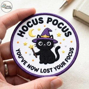 May include: A circular embroidered patch with a white background and purple border. It features a black cat wearing a purple witch hat, holding a yellow star wand. The text reads HOCUS POCUS YOU'VE NOW LOST YOUR FOCUS with yellow stars and a moon.