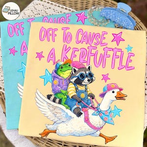 以下が含まれることがあります： 「OFF TO CAUSE A KERFUFFLE」のテキストが入った、2つのカラフルなグラフィックアートプリント。1つは水色、もう1つはベージュです。ベージュのプリントには、カエル、アライグマ、ガチョウのイラストが描かれています。青いヘアクリップとジュエリーが背景にあります。