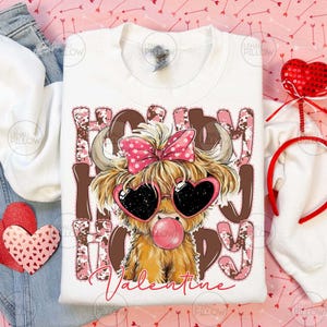 May include: White sweatshirt with a graphic of a Highland cow wearing heart-shaped sunglasses and a pink bow. The text "Happy Valentine" is printed on the shirt in pink and brown. A Valentine's Day themed design.