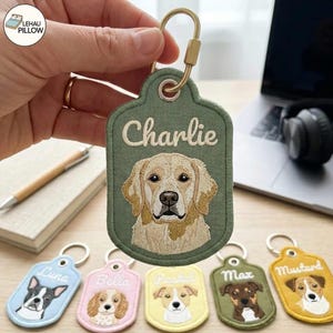Pode incluir: Um porta-chaves bordado verde-azeitona com o retrato de um golden retriever e o nome Charlie bordado em fio branco. A etiqueta tem um mosquetão de metal dourado preso a um ilhó reforçado no topo do tecido.
