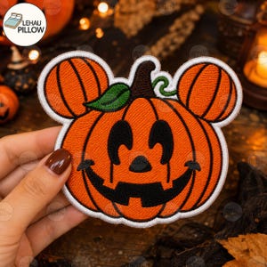 Może przedstawiać: Haftowana naszywka w kształcie dyni z dwoma mniejszymi okrągłymi uszami, czarną wyszywaną twarzą jack-o'-lantern, brązową łodygą i zielonym liściem, całość obwiedziona grubą białą ramką na ciemnym tle.
