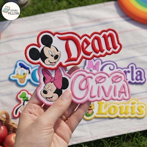 Peut inclure: Un écusson brodé personnalisé représentant Minnie Mouse avec un nœud rose et le prénom Olivia en lettres roses, bordé de rouge. D'autres écussons sont visibles, notamment le prénom Dean avec Mickey Mouse, Donald Duck, Goofy et Louis.