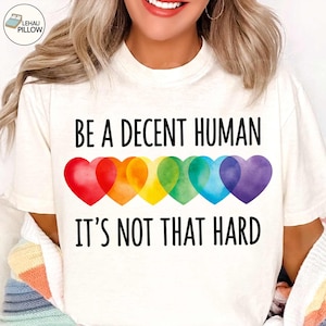 May include: A cream-colored t-shirt featuring the black text BE A DECENT HUMAN IT'S NOT THAT HARD. Between the two lines of text is a horizontal row of six overlapping watercolor-style hearts in red, orange, yellow, green, blue, and purple.