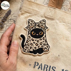 Puede incluir: Un parche bordado que muestra un gato negro con una capa con capucha de estampado de leopardo y un lazo a juego. El gato tiene grandes ojos color ámbar y una pequeña nariz rosa. El parche está sujeto a una bolsa de lona beige con la palabra PARIS.