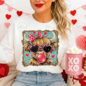 Peut inclure: Sweat-shirt blanc avec un dessin de vache des Highlands portant des lunettes de soleil en forme de c&oelig;ur et un n&oelig;ud rouge. La vache souffle une bulle de chewing-gum. Le motif est encadr&eacute; d'une bordure l&eacute;opard et de petits c&oelig;urs rouges.