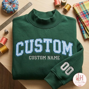Peut inclure: Un sweat-shirt vert foncé avec le mot CUSTOM en lettres bloc à carreaux vichy bleu clair et blanc, et le texte CUSTOM NAME brodé en blanc en dessous. La manche affiche le nombre 00 en blanc, avec une règle métallique visible à proximité.