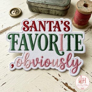 May include: A white embroidered patch with a scalloped edge featuring the text SANTA'S in red, FAVORITE in dark green with a pink letter I, and the word obviously in a cursive pink font, all stitched onto a white fabric base.