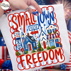 Könnte beinhalten: Weißes Kissen mit einem patriotischen Design mit den Worten "Small Town Freedom" in Rot und Blau. Das Design umfasst Illustrationen eines Wasserturms, Häusern und einer Kirche mit einem blau-weiß gestreiften Hintergrund.