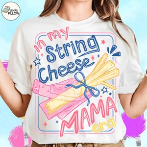 Puede incluir: Camiseta blanca con un diseño gráfico colorido. El diseño presenta el texto "In my String Cheese Mama Era" en rosa, azul y amarillo, junto con una ilustración de queso en hebras. La camiseta tiene cuello redondo y mangas cortas.