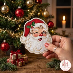 Könnte beinhalten: Ein gestickter Aufnäher mit dem Gesicht eines zwinkernden Weihnachtsmanns, einer roten Mütze, einem weißen Bart und grünen Stechpalmenblättern. Das Design ist mit weißem, rotem und grünem Garn und einer detaillierten Umrandung gefertigt.