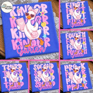 May include: A blue t-shirt featuring a white duck wearing a pink bow and heart-shaped sunglasses, carrying a pink notebook and bag. The design includes colourful stars, apples, and text indicating various school grades like Kindergarten and First Grade.