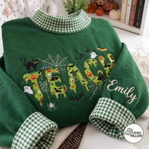 May include: A dark green sweatshirt featuring a high collar and cuffs with a white and green gingham pattern. The front displays the word TEACH in large, light green letters filled with Halloween motifs, surrounded by embroidered ghosts, bats, and spiderwebs. The name Emily is embroidered in white script on the sleeve.