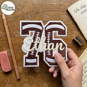 May include: A brown and white embroidered patch in the shape of the number 76, featuring a football-themed design with laces. The name Ethan is stitched in white cursive script across the center of the numbers, creating a personalized sports-themed applique.