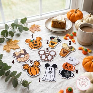 May include: A collection of eight embroidered patches featuring autumn and Disney-inspired designs, including a pumpkin, a beverage cup, a donut, a pretzel, and a ghost, each labeled with a number from one to eight on a white fabric surface.