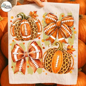 May include: A white fabric towel featuring a repeating graphic print of two pumpkins and two bows. The pumpkins have orange and brown leopard print sections with white football laces. The orange bows also feature white football lace patterns.