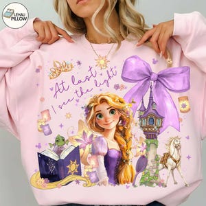 May include: Pink sweatshirt featuring a Rapunzel graphic with the text "At last I see the light." The design includes a tower, horse, and a large purple bow. The sweatshirt has a crew neck and long sleeves.