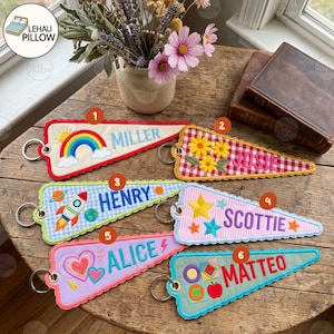 May include: Six triangular fabric pennant keychains with metal rings, each featuring embroidered names and motifs: a rainbow for Miller, flowers for Josie, a rocket for Henry, stars for Scottie, hearts for Alice, and geometric shapes for Matteo.