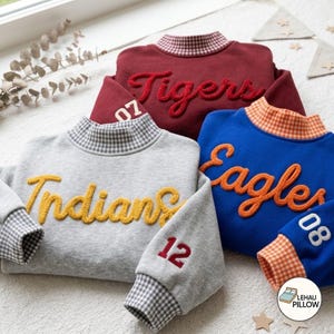 May include: Three long-sleeve sweatshirts in gray, maroon, and blue. The gray sweatshirt features yellow text reading Indians and the number 12. The maroon sweatshirt has red text reading Tigers and the number 07. The blue sweatshirt has orange text reading Eagles and the number 08. Each has a gingham collar and cuffs.