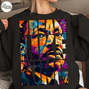 May include: Black sweatshirt featuring a vibrant, multi-colored graphic portrait with the text "DREAM LIKE KING". The artwork uses a collage style with bold colors like orange, purple, and blue, creating a striking visual.