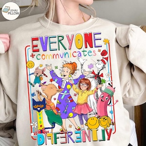 Puede incluir: Sudadera beige con un gráfico colorido. El diseño presenta el texto "EVERYONE COMMUNICATES DIFFERENTLY", personajes de dibujos animados y piezas de rompecabezas. La sudadera es de cuello redondo.