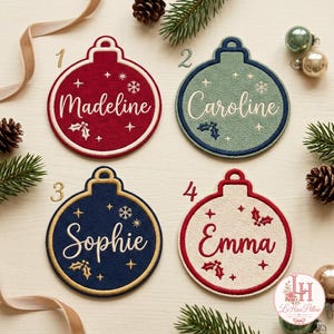 May include: Four circular embroidered ornaments featuring the names Madeline, Caroline, Sophie, and Emma. Each ornament displays a unique color scheme with stitched snowflake and holly motifs, numbered one through four in gold or red text.