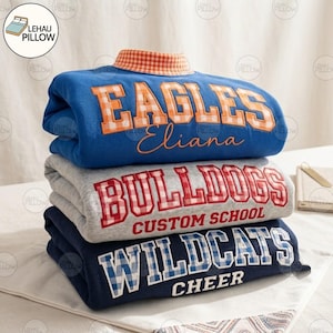 May include: A stack of three folded sweatshirts. The top is blue with orange gingham lettering reading EAGLES and Eliana. The middle is grey with red gingham lettering reading BULLDOGS CUSTOM SCHOOL. The bottom is navy with blue gingham lettering reading WILDCATS CHEER.