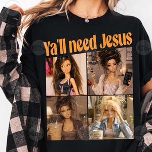 May include: A black t-shirt featuring the orange text "YA'LL need Jesus" above a four-panel collage of fashion dolls. The dolls are depicted in various settings, including holding a makeup brush, a smartphone, a coffee cup, and a mug.