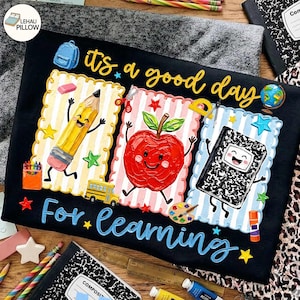May include: A black fabric item featuring the text 'it's a good day For learning' in yellow and blue. The design includes three scalloped frames containing a cartoon pencil, a red apple, and a composition notebook, plus a school bus and globe.