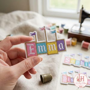 Peut inclure: Un écusson brodé rectangulaire affichant le prénom Emma en lettres blanches sur quatre blocs colorés. Chaque bloc est surmonté d'un petit drapeau. L'écusson possède une bordure blanche et est présenté sur un fond neutre avec du matériel de couture.