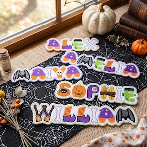 May include: Four custom name signs featuring Halloween-themed letters. The signs spell out ALEX, AMELIA, MAIYA, SOPHIE, and WILLIAM, decorated with motifs like pumpkins, bats, witches' hats, bones, spiderwebs, and Frankenstein-style bolts.
