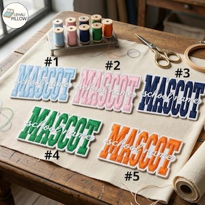Könnte beinhalten: Fünf gestickte Aufnäher mit dem Wort MASCOT in Blockbuchstaben und dem Text school name in Schreibschrift. Die Aufnäher sind hellblau, rosa, dunkelblau, grün und orange. Ein Metallgarnrollenhalter, eine Schere und Stoff sind zu sehen.