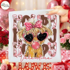 May include: White square pillow with a cartoon Highland cow wearing heart-shaped sunglasses and a pink bow. The words "Happy Valentine" are printed in brown and pink. The cow is blowing a pink bubble.