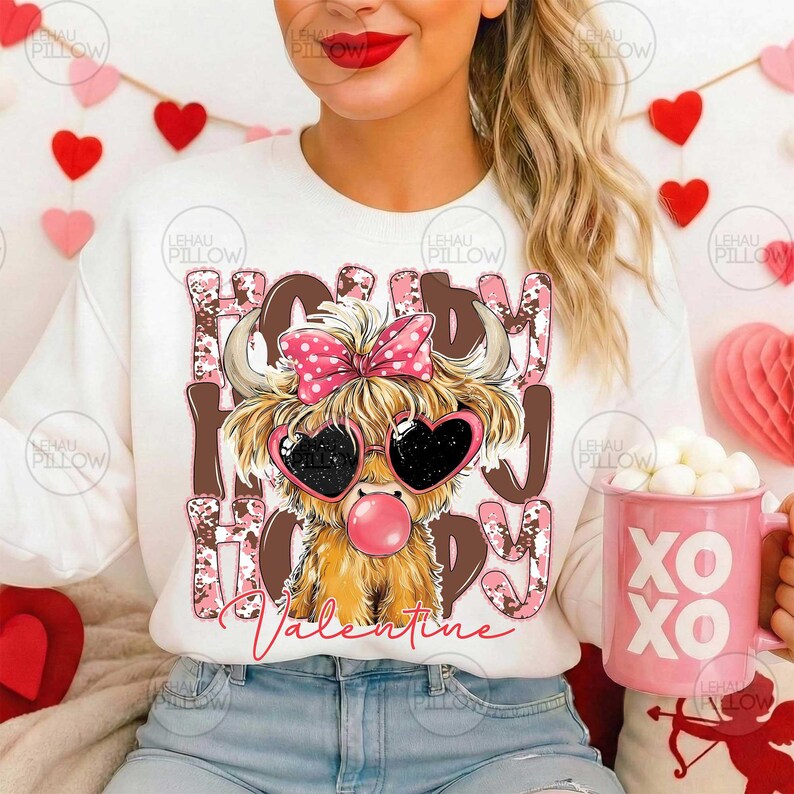 May include: White sweatshirt with a graphic of a Highland cow wearing heart-shaped sunglasses and a pink bow. The text "Happy Valentine" is printed on the shirt. A pink mug with "XO XO" is visible.