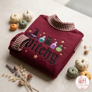 May include: A maroon sweatshirt featuring a brown and white gingham collar and cuffs. The front displays embroidered icons of a witch hat, a black cat, and three potion bottles, with the word "Witchy" in black script and small yellow stars.