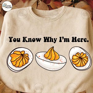 Op de afbeelding: Beige sweatshirt met de tekst "You Know Why I'm Here." en illustraties van gevulde eieren. De eieren hebben een witte basis en zijn bedekt met een gele vulling.